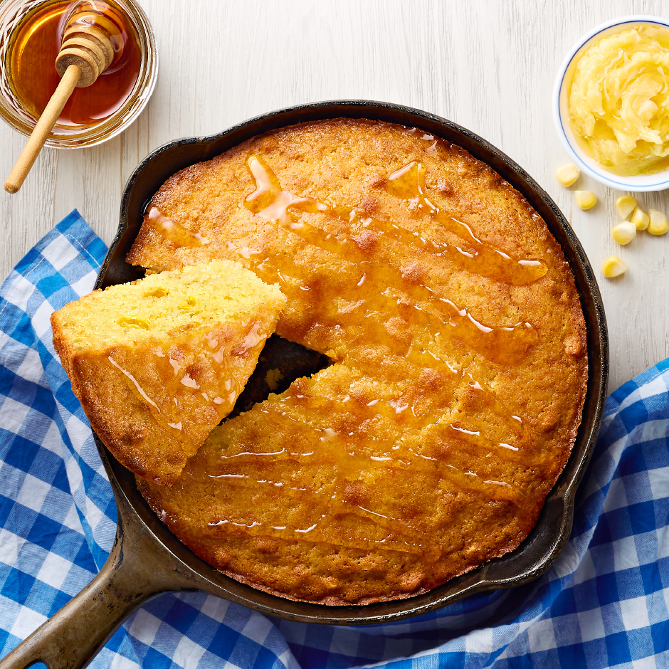 A cast-iron pan of Honey Flavored Cornbread on a blue-and-white gingham napkin.