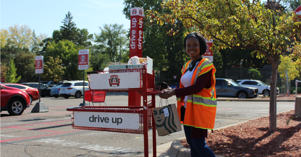 A Drive Up team member holding a Starbucks bag poses with a cart containing a guest’s order in a parking lot.